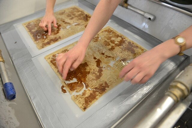 Das Foto zeigt ein mit Wasser und Faserbrei gefülltes Becken, in dem zwei historische beschädigte Dokumente liegen. Von zwei Personen sind die Hände zu sehen, mit denen abgebrochene Papierstücke an die richtige Stelle in den Dokumenten gebracht werden.