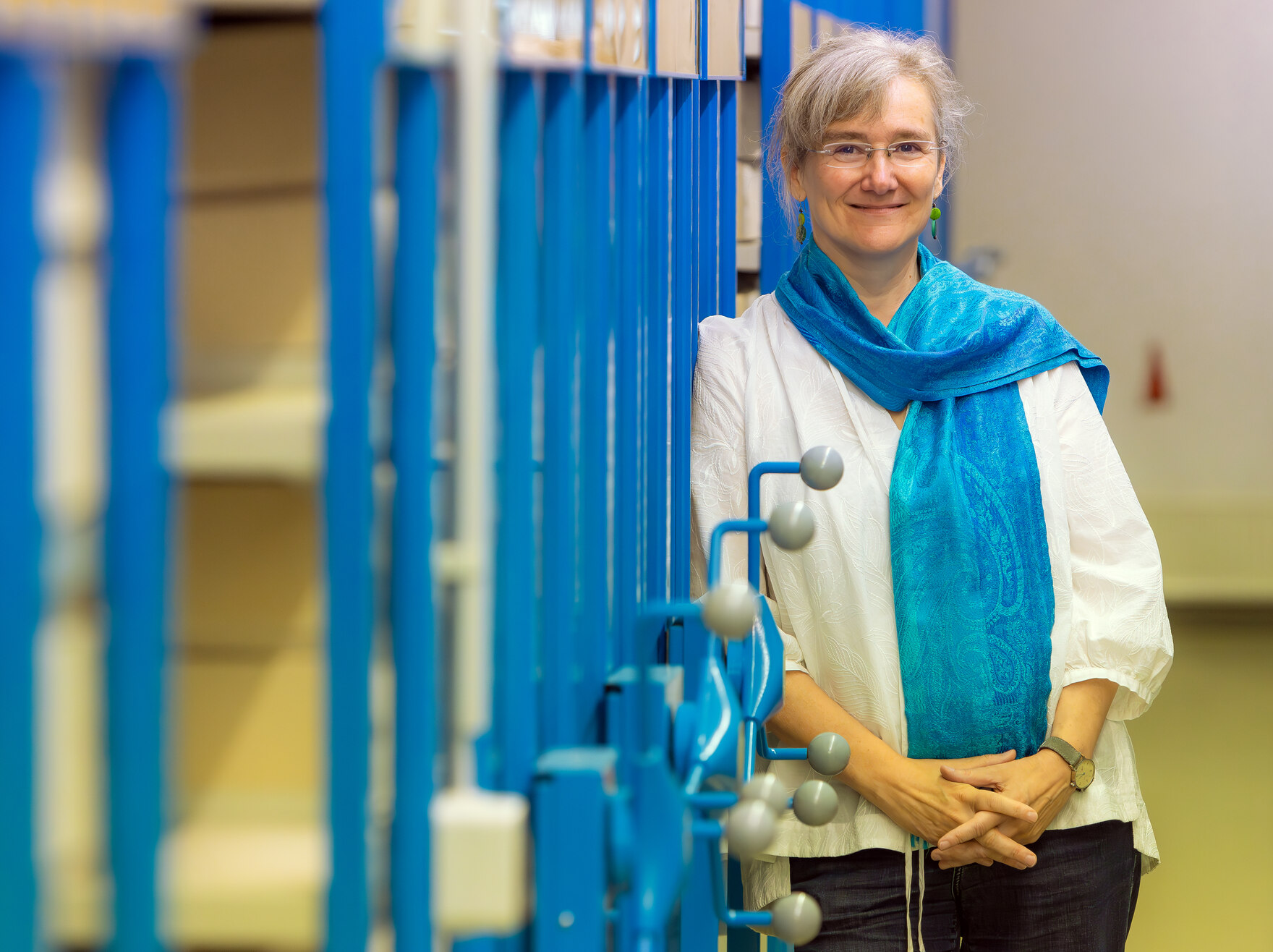 Das Foto zeigt links eine blaue Rollregalanlage, an der sich eine Frau anlehnt und lächelt. Sie trägt eine Brille, eine weiße Bluse und ein blaues Tuch darüber.